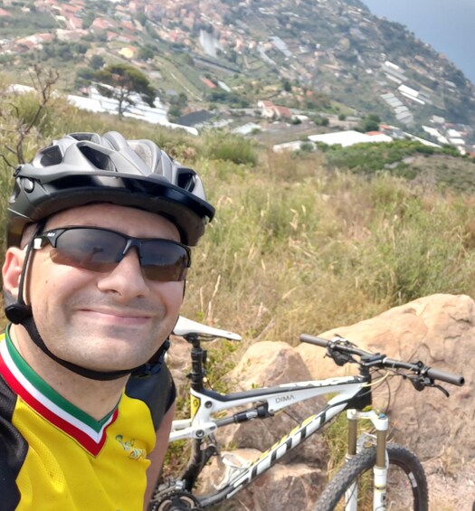 A person wearing a cycling helmet and a yellow jersey, standing next to a mountain bike with a rocky landscape in the background. 5