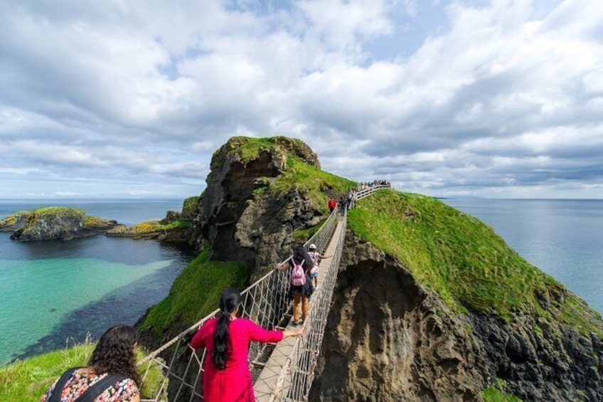 A coastal cliff with a rope bridge, clear blue water, and a grassy hill. 1
