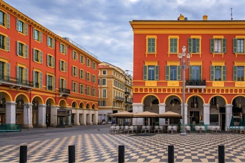 A square with checkered pavement, red and yellow buildings with green shutters, and outdoor tables and chairs. 3