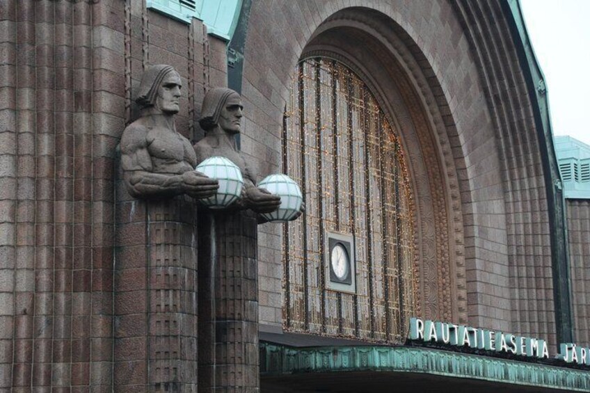 A building facade with statues of muscular figures holding globes, a clock, and the inscription 'RAUTATIEASEMA JAR'. 2