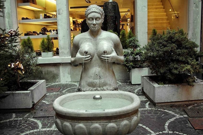 A statue of a woman with exposed breasts in a courtyard with a fountain, surrounded by potted plants and a shoe store in the background. 4