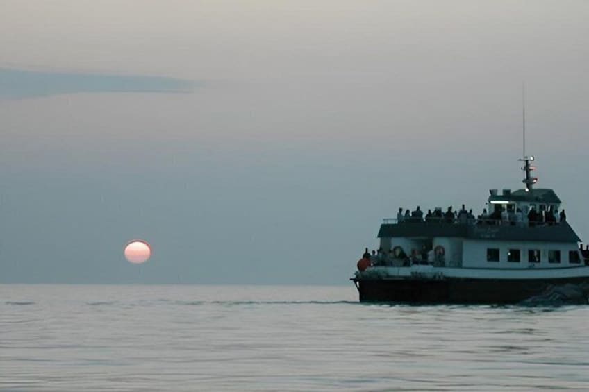 A boat with people on deck, a calm sea, and a setting sun. 1