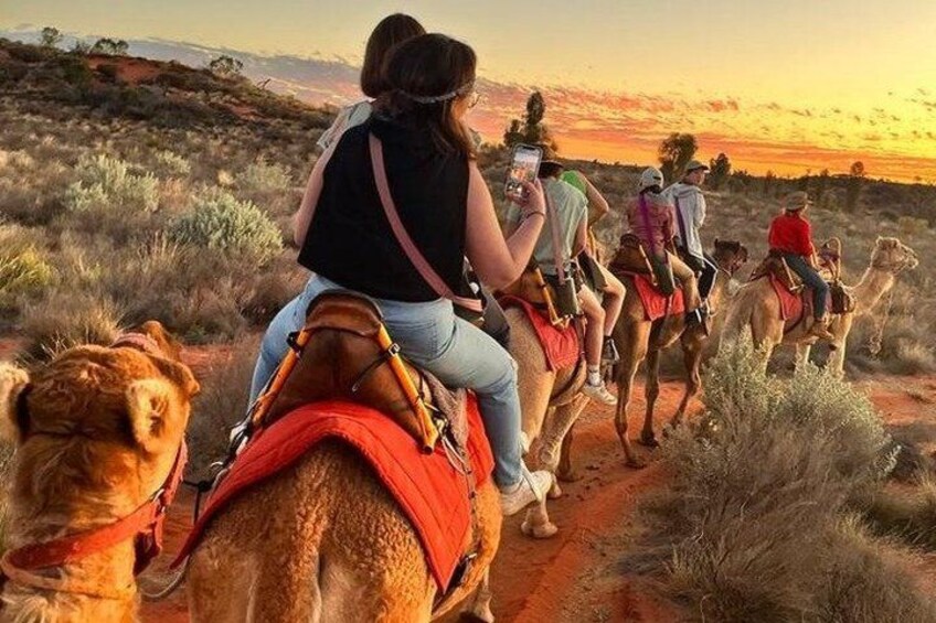 A group of people riding camels on a dirt path with a sunset in the background. 3