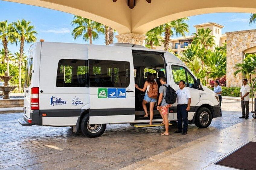 A white van with 'CABO ADVENTURES' and 'dolphins' logos, parked at a resort entrance with palm trees and a fountain in the background. 2
