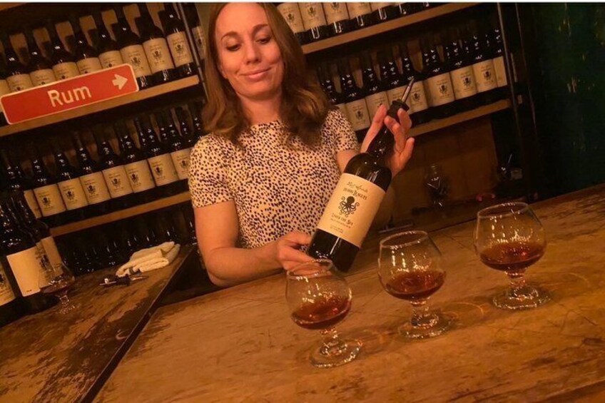 A person pouring a bottle of rum into glasses on a bar counter with shelves of bottles in the background. 4