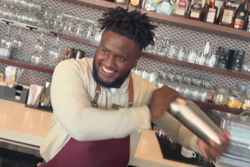 A person in a bar setting, wearing a white shirt and maroon apron, holding a shaker bottle. 1