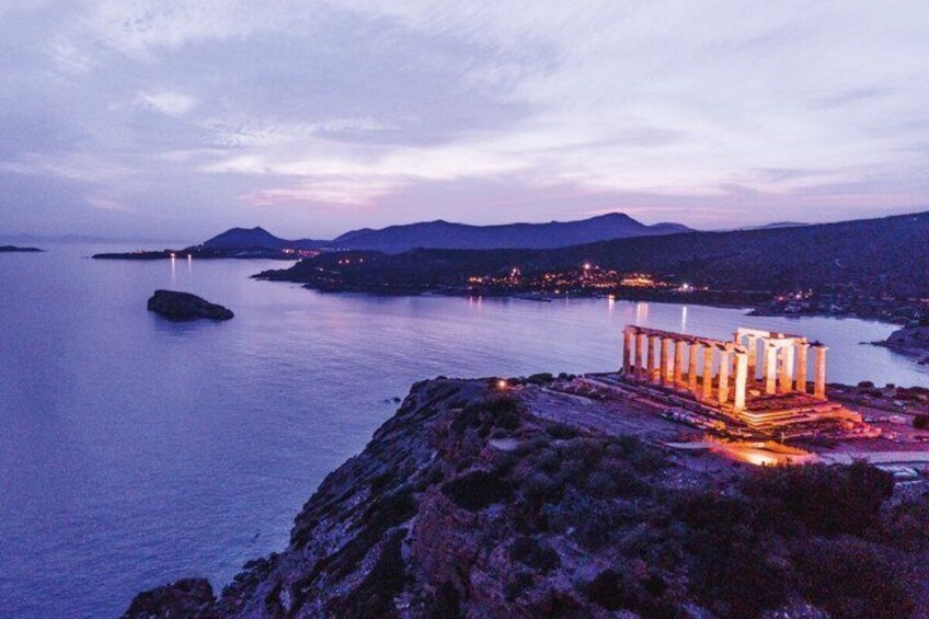 A coastal landscape with a prominent ancient Greek temple illuminated at dusk, a rocky shoreline, and distant mountains. 4
