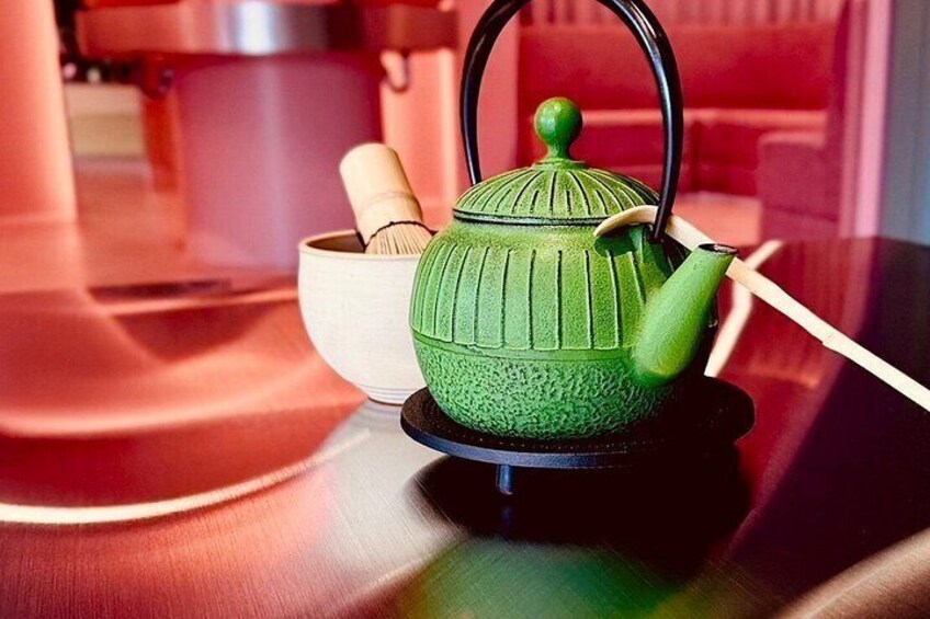 A green teapot with a bamboo handle on a black coaster, a white bowl with a bamboo whisk, and a red chair in the background. 1