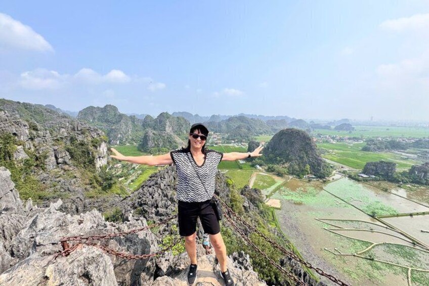 A person standing on a rocky outcrop with arms outstretched, overlooking a valley with terraced fields. 1