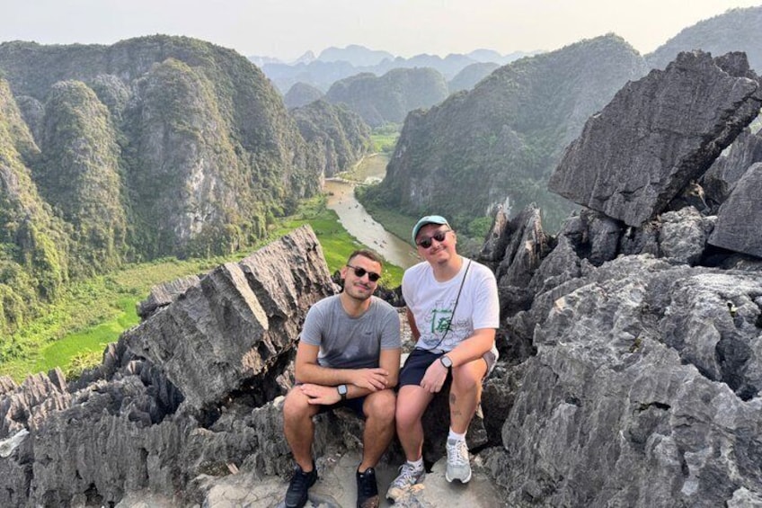 Two people sitting on a rocky ledge with a river and mountains in the background. 5
