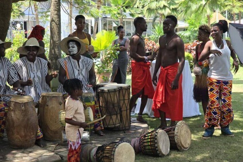 A group of people playing drums in an outdoor setting with trees and a building in the background. 1