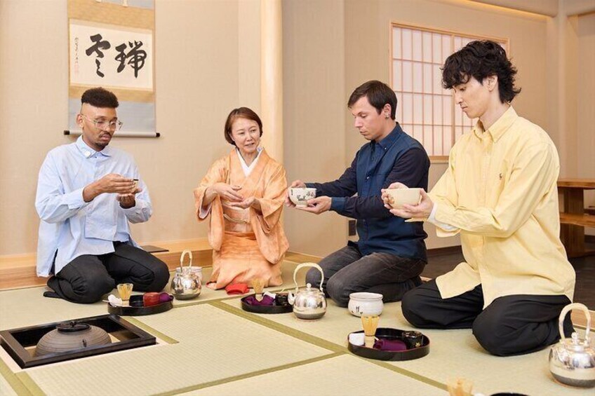 Four people sitting on tatami mats in a traditional Japanese room with tea sets. 4