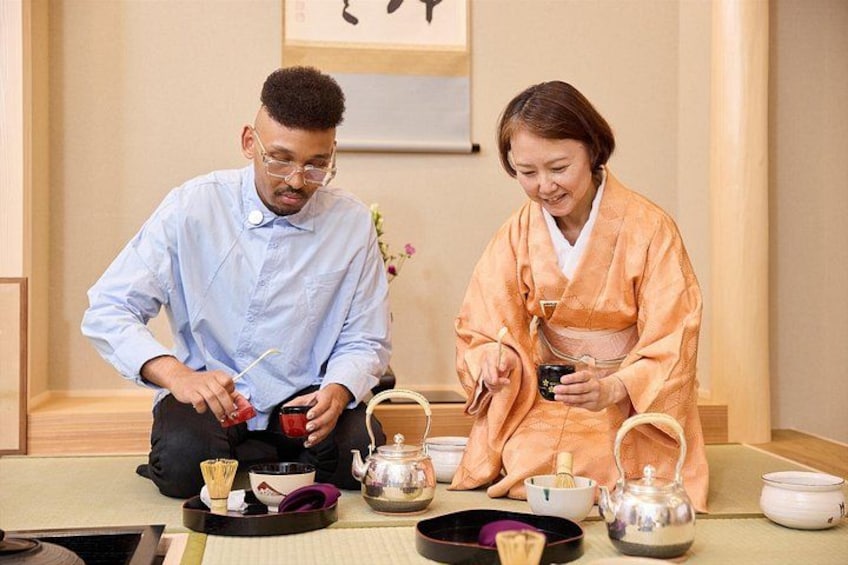 Two people in traditional attire sitting on tatami mats, with tea sets and a framed picture in the background. 5