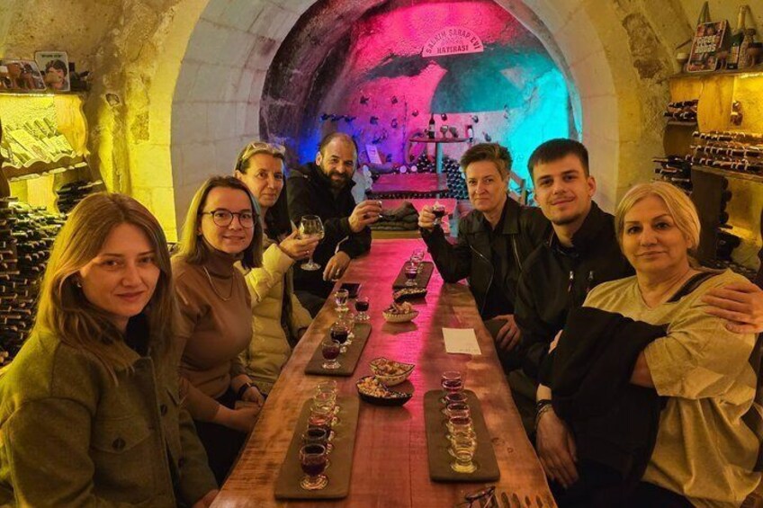 A group of people seated at a long table in a wine cellar with bottles and glasses. 3