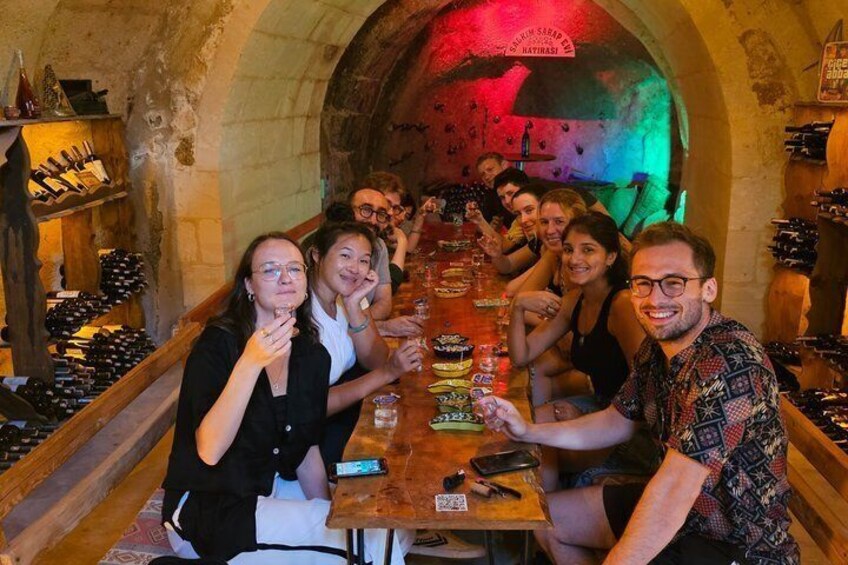 A group of people seated at a long table in a wine cellar with wine racks on the walls. 1