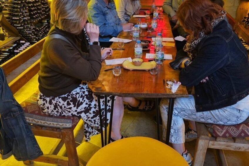 A group of people seated at a wooden table in a restaurant with bottles and glasses on it. 4