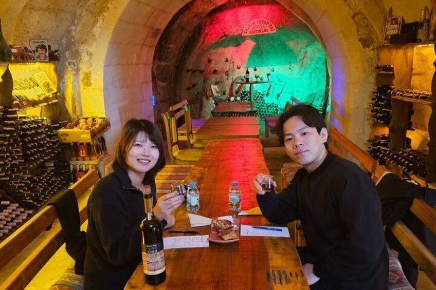 Two people seated at a table in a wine cellar with bottles and glasses. 2