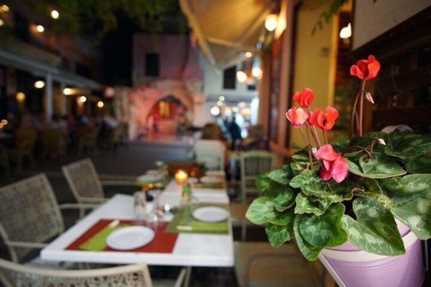 An outdoor dining area with tables set for two, a potted plant with red flowers, and a street in the background. 1