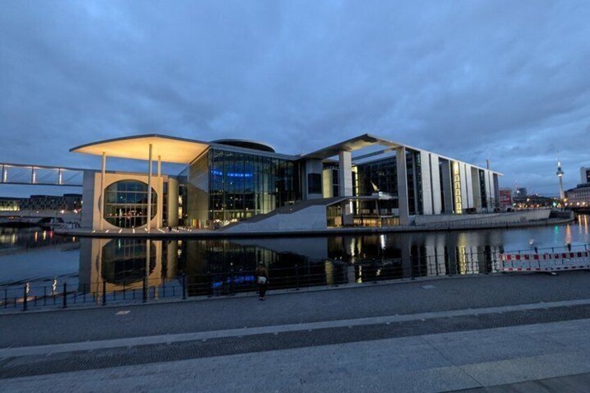 A modern architectural building with a curved glass facade, situated by a waterfront with a bridge in the background. 1