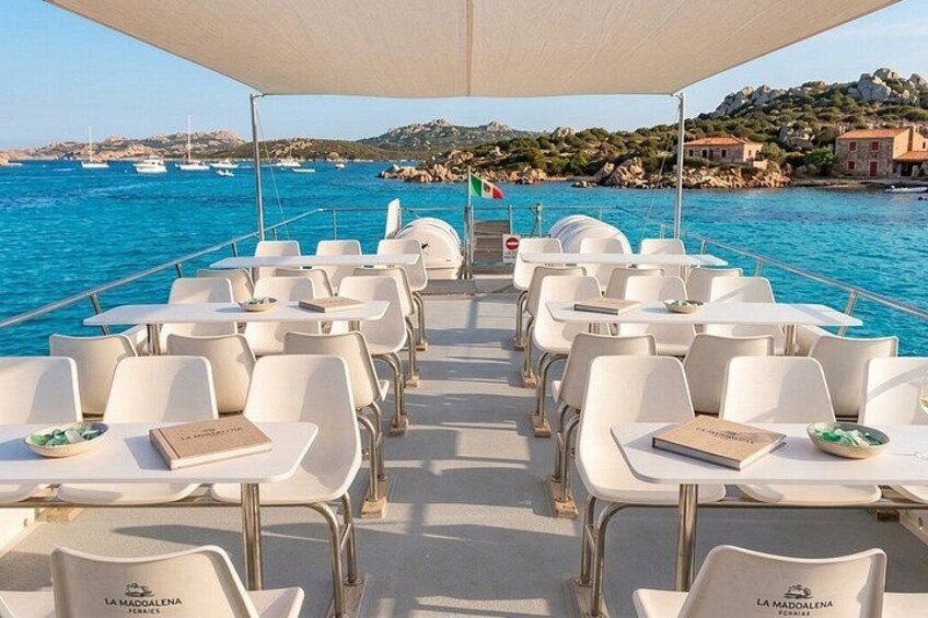 A boat deck with white tables and chairs, a flag, and a view of the sea and coastline. 2