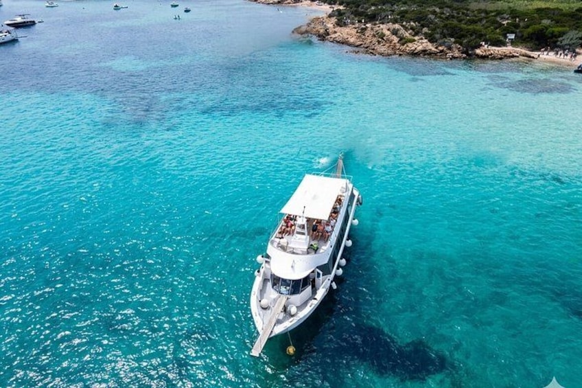 A white boat with a covered deck and multiple windows, anchored in clear turquoise water near a rocky shoreline with sparse vegetation. 1