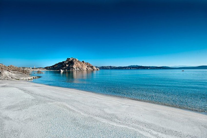 A beach with clear blue water, a rocky outcrop, and distant hills. 4