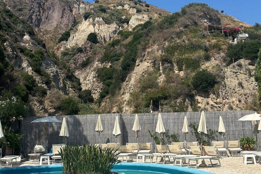 A pool with lounge chairs and umbrellas, a rocky hillside, and a building in the background. 5