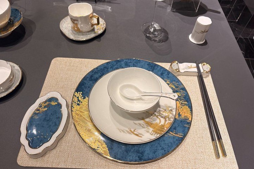 A table set with a blue and gold plate, a white bowl, a white cup, a glass, and a white candle. 3