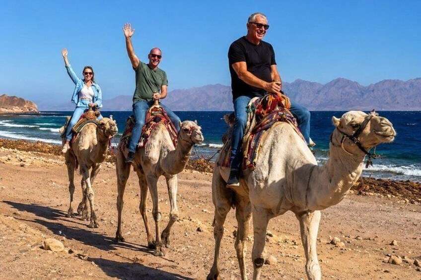 Three people riding camels on a beach with mountains in the background. 4
