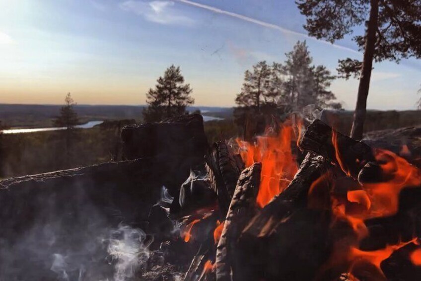 A campfire with visible flames and smoke, surrounded by trees and a distant view of a body of water. 2