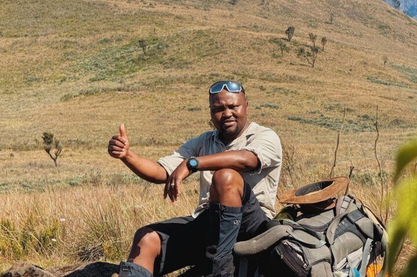 A person sitting on the ground with a backpack, surrounded by dry grass and hills. 1