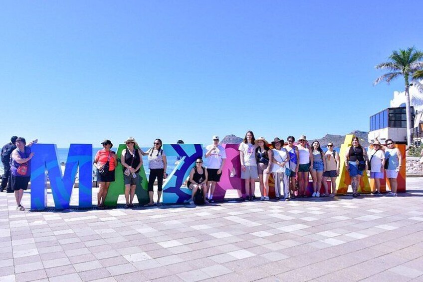 A group of people standing in front of large colorful letters spelling 'MEXICO' on a sunny day. 2