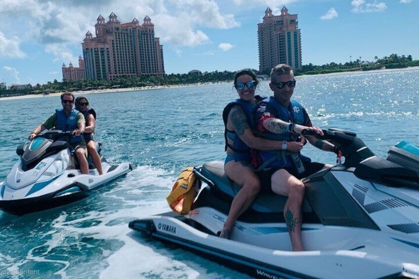 Two people on a jet ski and another on a water ski, with a resort and buildings in the background. 1
