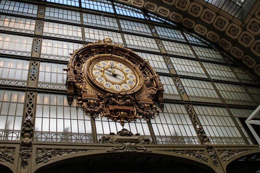 A large ornate clock with Roman numerals, set against a backdrop of a glass-paneled wall with intricate metalwork. 3