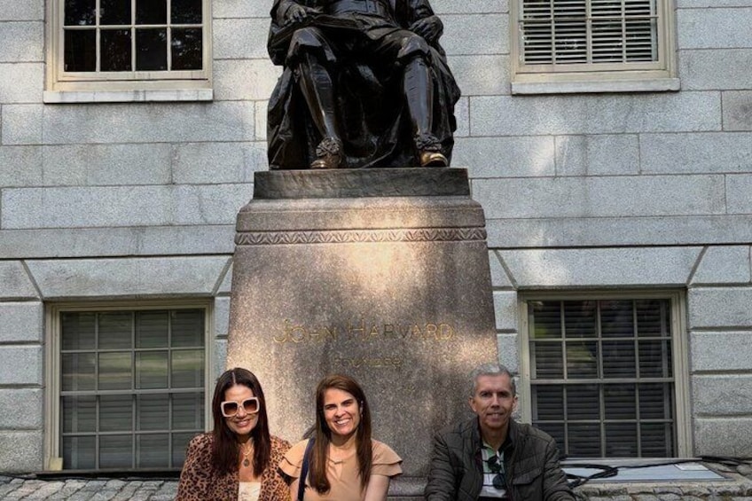 A statue of John Harvard on a pedestal with inscriptions, set against a building facade with windows and shutters. 1