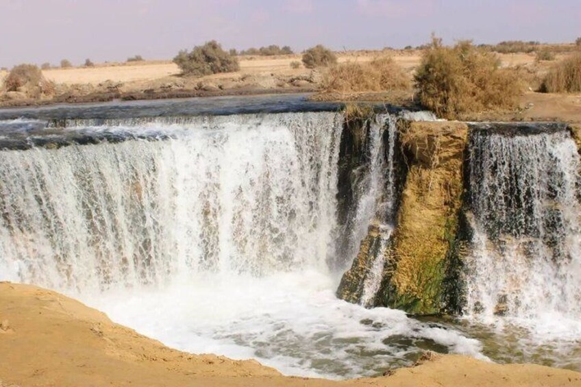 A waterfall cascading over a rocky ledge into a sandy area with sparse vegetation in the background. 4