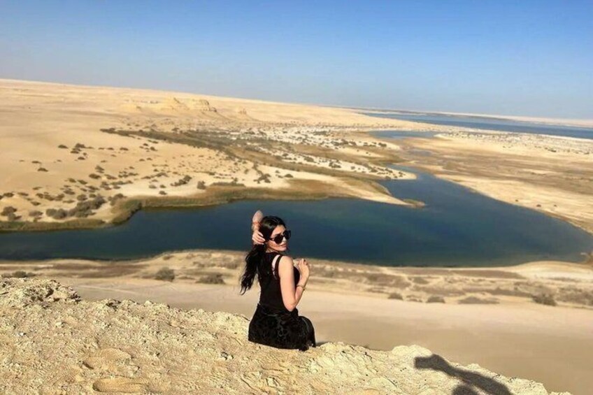 A person sitting on a sandy dune overlooking a desert landscape with a body of water. 1
