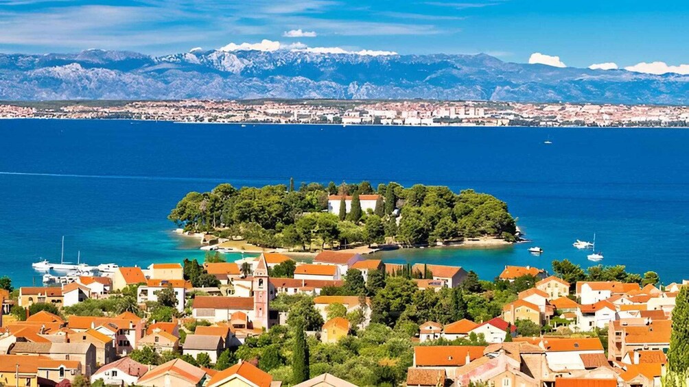 A coastal town with red-roofed buildings, a small island with trees, and a mountain range in the background. 1