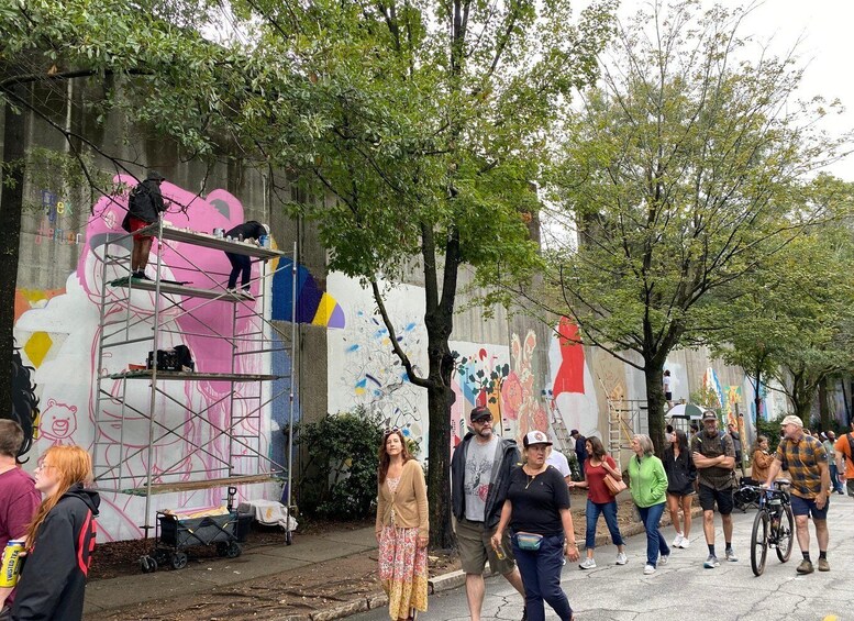 A street with a mural being painted on a wall, people walking by, and trees lining the sidewalk. 2