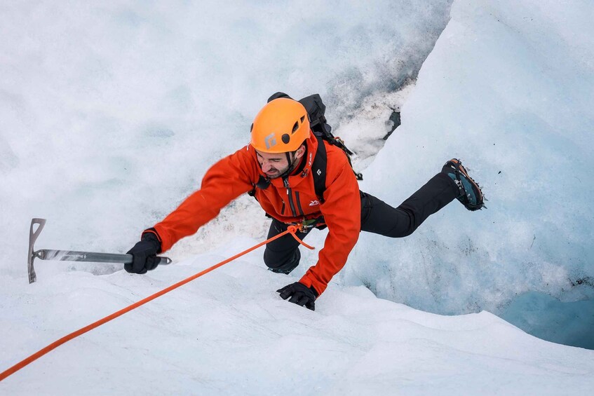 A person in an orange jacket and helmet using ice axes to climb a snowy slope. 1