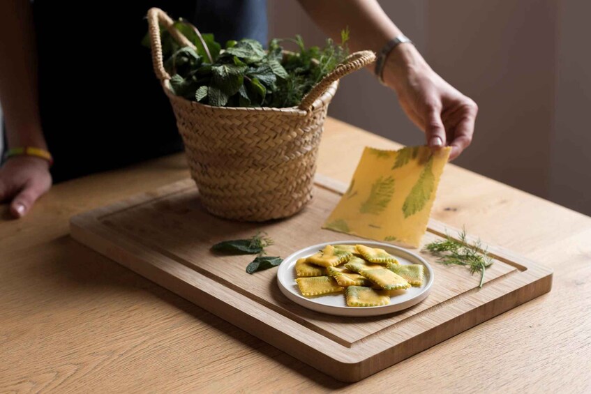 A wooden cutting board with a plate of ravioli, a woven basket with green leaves, and a hand holding a yellow envelope. 5