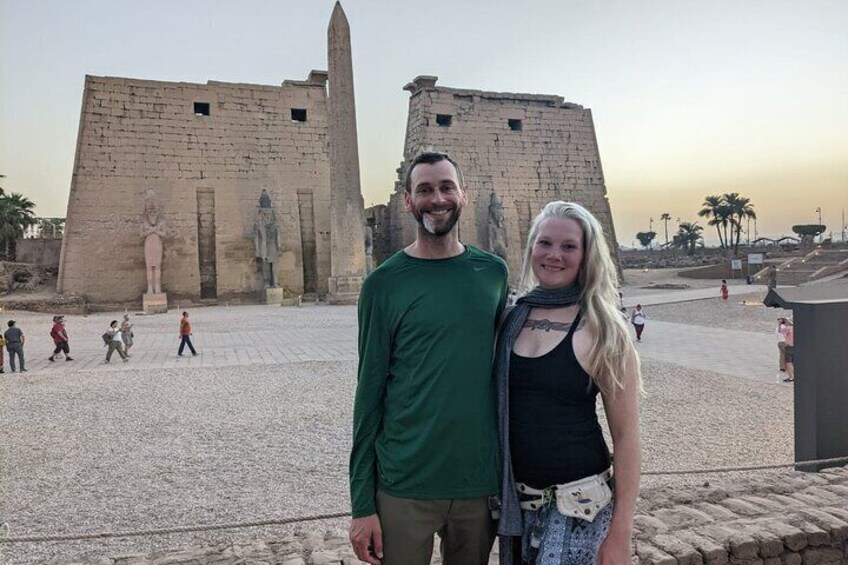 A couple standing in front of ancient Egyptian ruins with large statues and a tall obelisk. 1