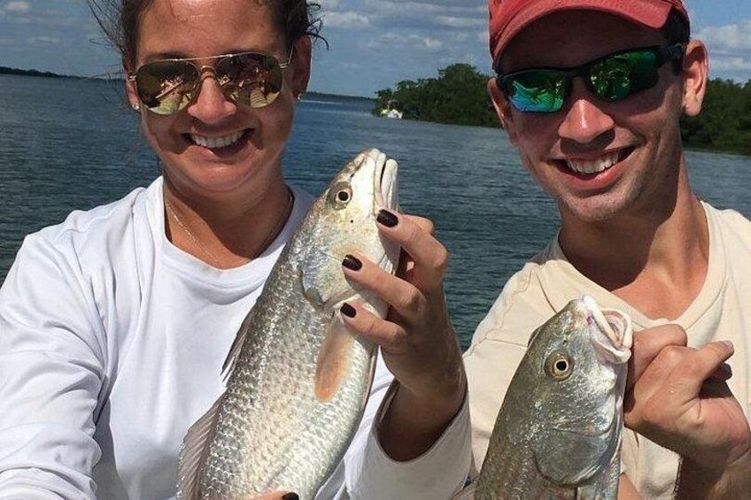 Two people holding fish by a body of water with a distant shoreline. 2