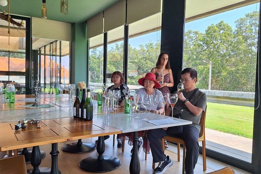 A group of people seated at a table with wine bottles and glasses, in a room with large windows and a view of greenery outside. 1