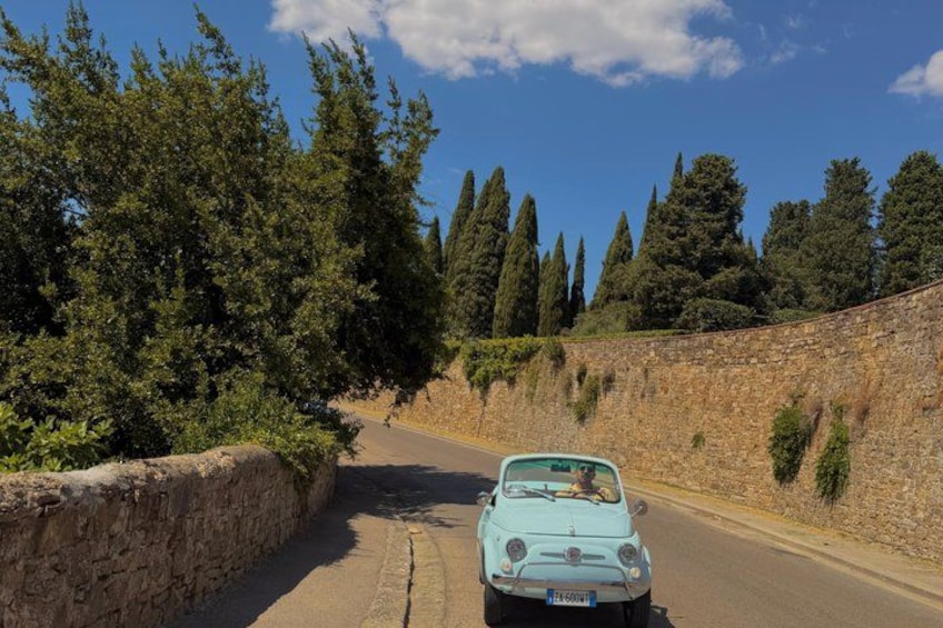A vintage car driving on a narrow road flanked by stone walls and dense trees. 2