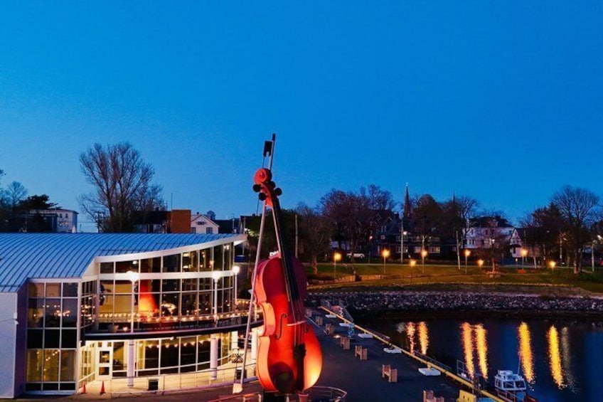 A building with a glass facade, a large cello sculpture, and a body of water reflecting lights. 1