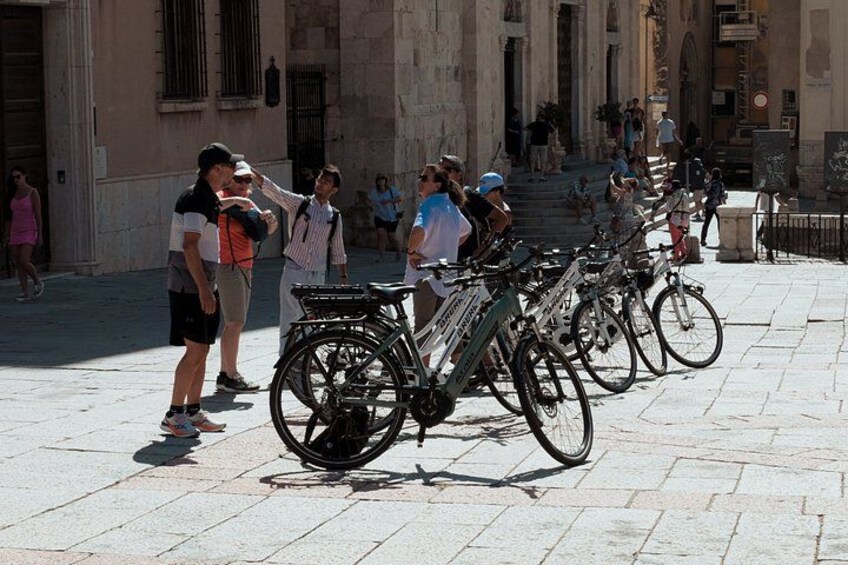 A row of bicycles parked on a cobblestone street, a building with columns, and people walking. 5
