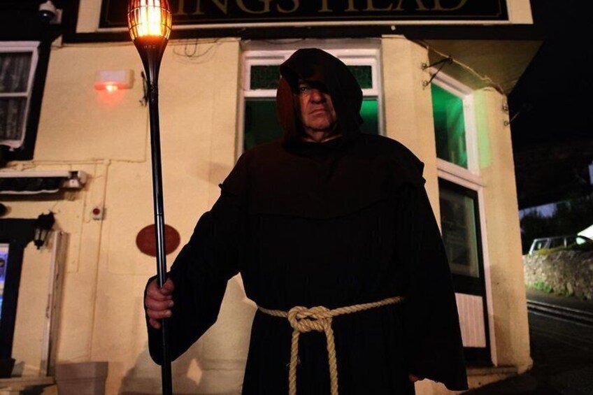 A person in a dark robe holding a torch in front of a building with a sign that reads 'KING'S HEAD.' 1