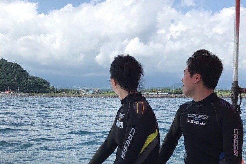 Two people in wetsuits on a boat, with a body of water and a distant landmass under a cloudy sky. 3