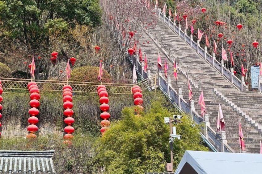 A staircase with red lanterns and flags, surrounded by greenery and trees. 4